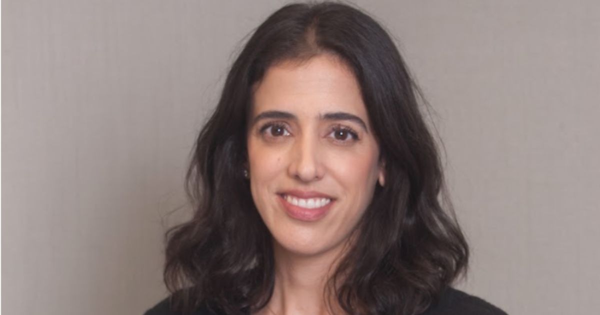 Mulher sorridente de cabelo castanho escuro ondulado, com camisola preta, em frente a um fundo neutro.