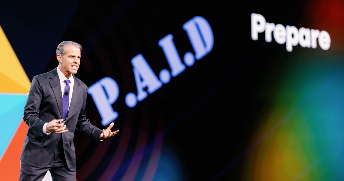 Imagem de Eric O'Neil, especialista em cibersegurança e ex-operador do FBI, durante sua palestra no evento VeeamON 2025. Ele está de terno escuro e gravata roxa, gesticulando enquanto fala no palco. Ao fundo, vê-se uma tela com a palavra "P.A.I.D" em destaque e o termo "Prepare" parcialmente visível, indicando o conteúdo de sua apresentação (hacker)