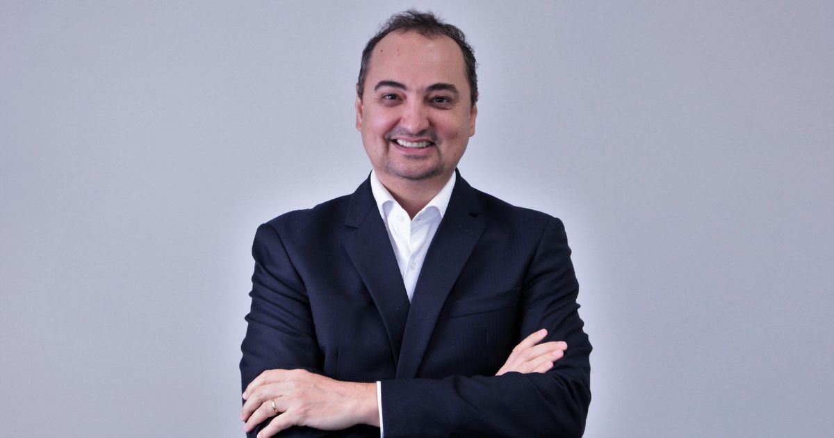 Homem de fato escuro e camisa branca sorridente, de braços cruzados, posando contra fundo cinzento.