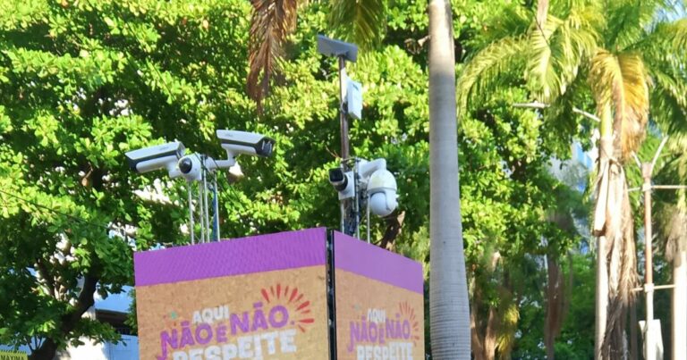 Imagem de um totem de segurança pública instalado em área externa com várias câmeras de vigilância voltadas em diferentes direções. O totem exibe o aviso colorido: 'AQUI NÃO É NÃO. RESPEITE', indicando uma campanha de conscientização contra o assédio, possivelmente em evento público. Ao fundo, há árvores tropicais e postes, sugerindo um ambiente urbano arborizado.