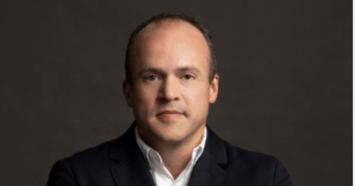 Um homem de pele clara, com cabelos curtos e ligeiramente recuados, está posando para um retrato profissional. Ele veste um blazer preto sobre uma camisa branca e calças bege. Seus braços estão cruzados, e sua expressão é séria e confiante. O fundo é escuro e neutro, destacando o sujeito na imagem (Intelbras)