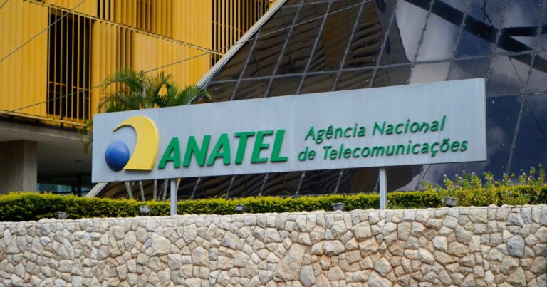 Placa da ANATEL (Agência Nacional de Telecomunicações) em destaque, com logotipo em azul, amarelo e verde, e o nome da instituição em letras verdes sobre um fundo metálico. Ao fundo, a fachada moderna do prédio com vidros espelhados e detalhes em amarelo. O cenário inclui vegetação e um muro de pedras na base da placa