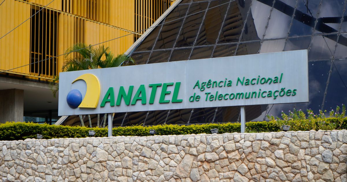 Placa da ANATEL (Agência Nacional de Telecomunicações) em destaque, com logotipo em azul, amarelo e verde, e o nome da instituição em letras verdes sobre um fundo metálico. Ao fundo, a fachada moderna do prédio com vidros espelhados e detalhes em amarelo. O cenário inclui vegetação e um muro de pedras na base da placa