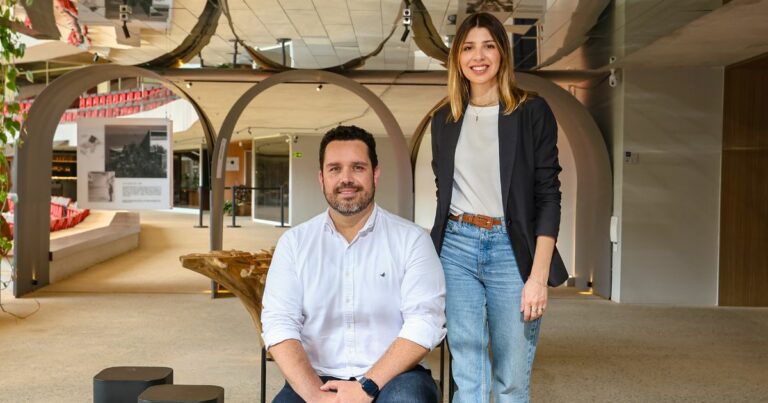 Um homem de barba curta, cabelo escuro e camisa branca está sentado, enquanto uma mulher de cabelo liso e loiro, vestindo um blazer preto e jeans, está de pé ao seu lado. Eles estão em um ambiente moderno, com arcos e espelhos no teto, dando um toque sofisticado ao local. Ao fundo, há uma exposição de fotografias e cadeiras vermelhas organizadas em arquibancadas (ecossistema)