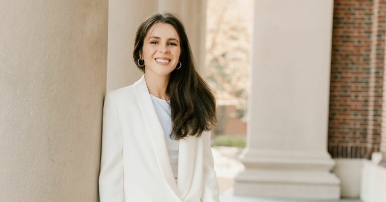 Mulher jovem a sorrir, vestida com blazer branco, encostada a uma coluna de pedra num espaço exterior com arquitetura clássica ao fundo.
