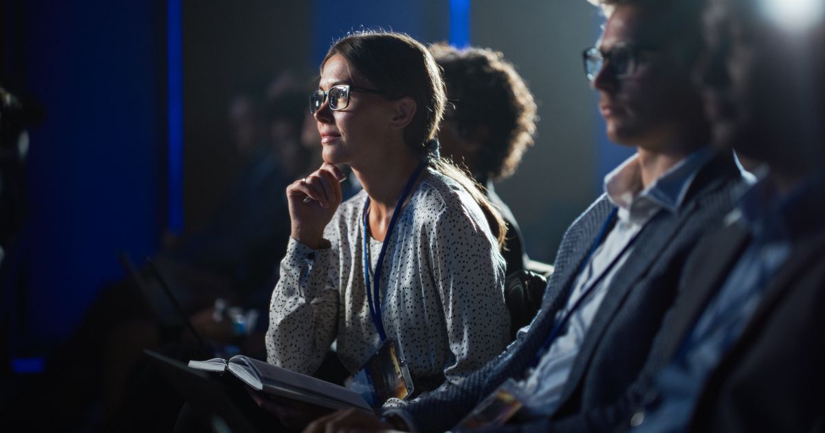 Mulher sentada com a mão no queixo, com caderno no colo e olhando para a frente com rosto de interesse e admiração