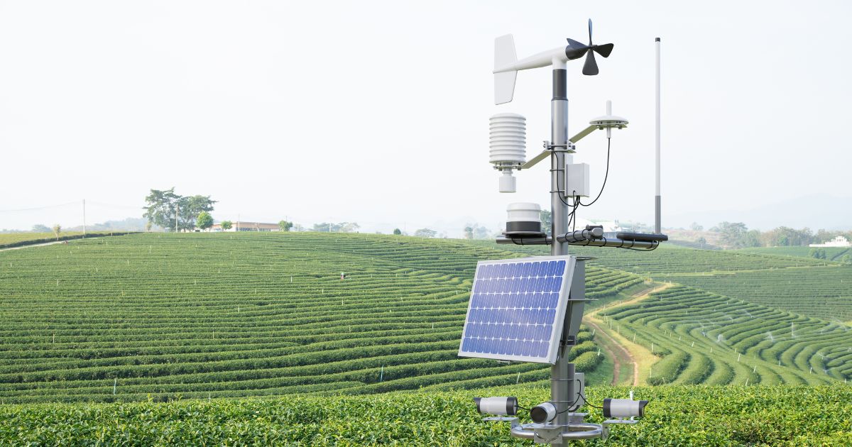 "Imagem de uma estação meteorológica moderna instalada em uma plantação. O equipamento conta com sensores, antenas, um painel solar e anemômetro, utilizados para coletar dados climáticos e ambientais. Ao fundo, vê-se uma plantação em grandes curvas e fileiras verdes, com sistema de irrigação visível e paisagem rural ao longe."