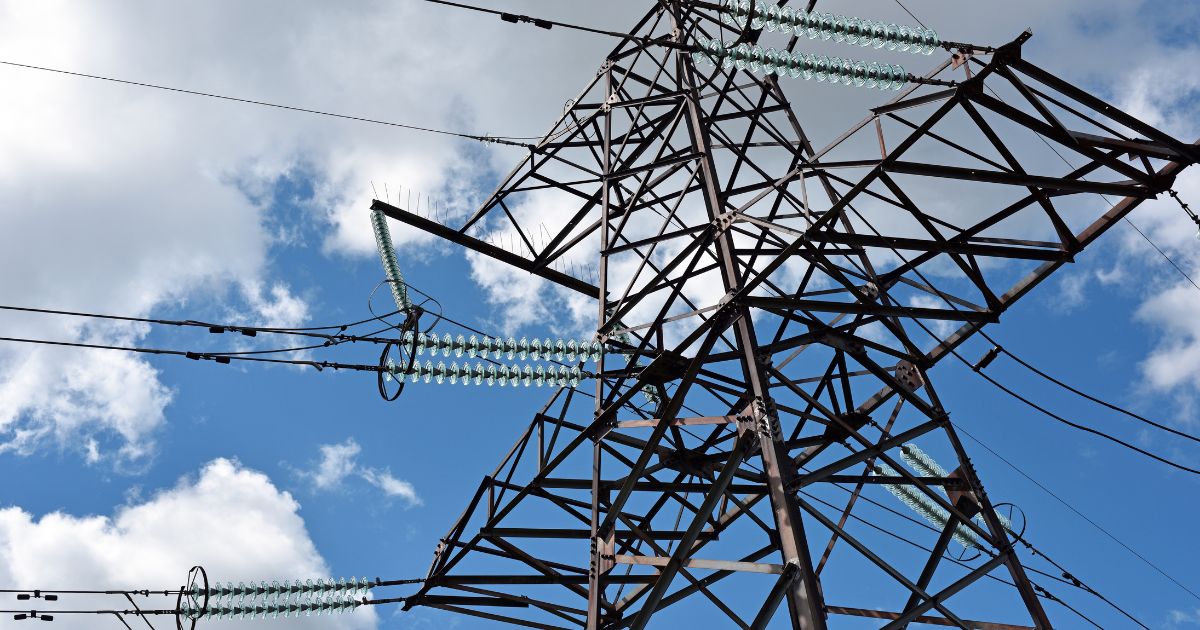 Fotografia de uma torre metálica de transmissão de energia elétrica vista de baixo contra um céu azul com algumas nuvens brancas. A estrutura de ferro é composta por uma malha de vigas diagonais e verticais, suportando cabos de alta tensão conectados através de isoladores de vidro verde-azulado em série.