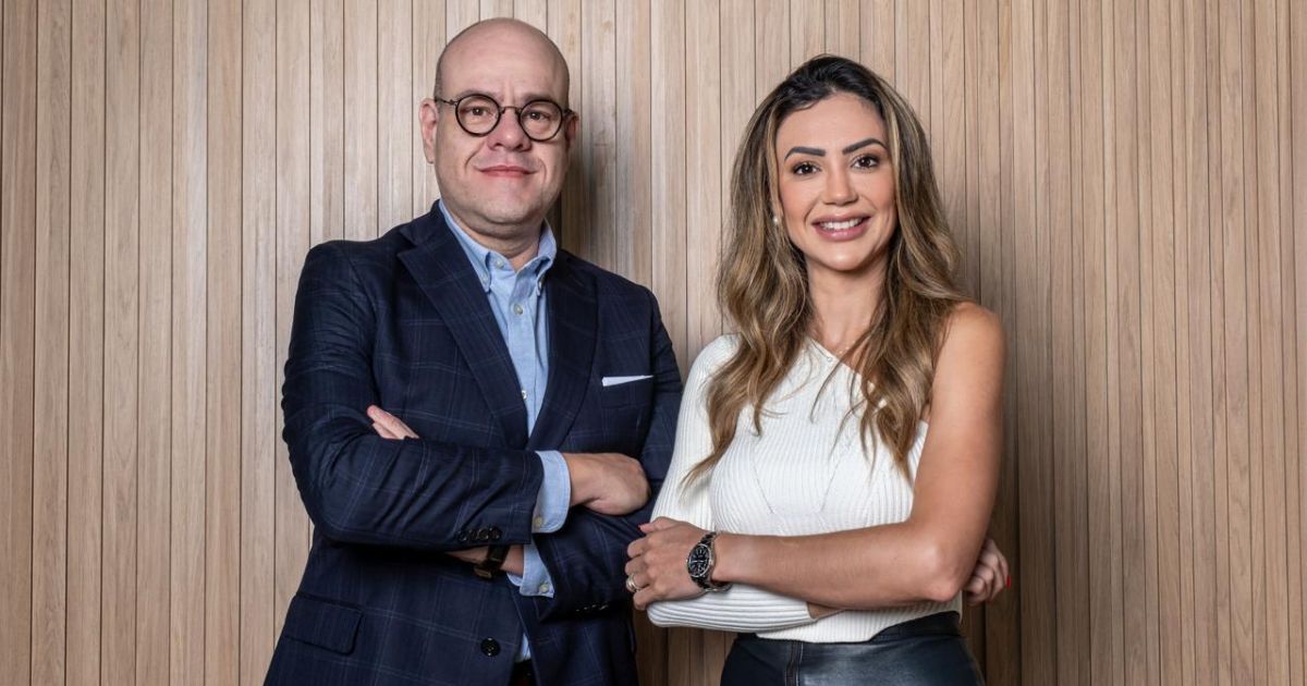 Shandrus Carvalho, vice-presidente da Virtueyes, e Taize Wessner, presidente da Virtueyes, posam lado a lado em frente a uma parede de madeira com ripas verticais. Shandrus usa terno escuro com camisa azul clara e óculos redondos, enquanto Taize veste uma blusa branca sem mangas e calça de couro preta, ambos com os braços cruzados e sorrindo