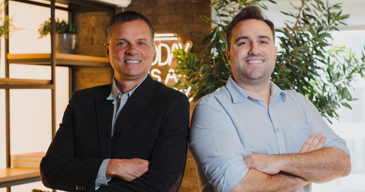 Pedro Chiamulera, CSO, e Francesco Weiss, novo CEO da Neotrust, posam sorrindo lado a lado com os braços cruzados. Pedro Chiamulera, à esquerda, veste blazer preto e camisa azul clara. Francesco Weiss, à direita, veste camisa social azul clara. Ao fundo, ambiente interno com plantas, estante decorada e letreiro luminoso desfocado