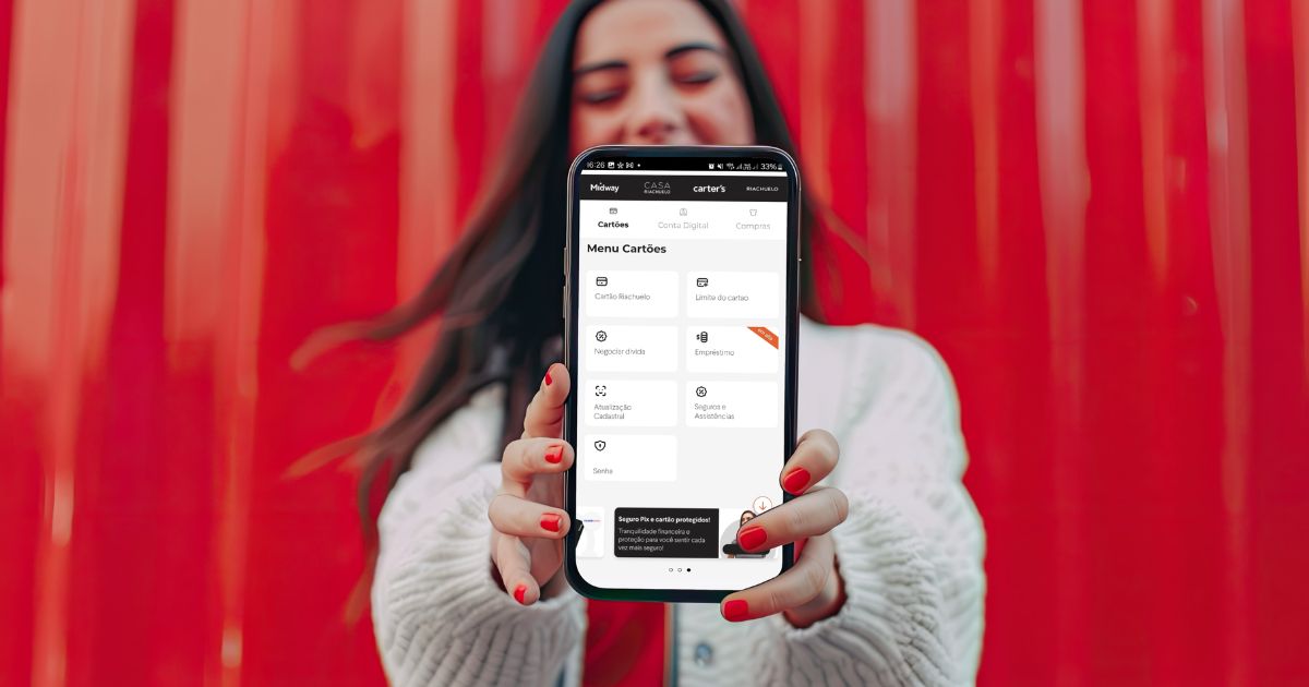 Mulher sorridente com unhas pintadas de vermelho, vestindo suéter branco, segura um celular em destaque, exibindo a tela do aplicativo da Riachuelo. Ao fundo, uma parede vermelha desfocada. A interface do app mostra o menu de cartões com opções como cartão Riachuelo, limite do cartão, negociação de dívidas, empréstimo, atualização cadastral, seguros e assistências, e senha