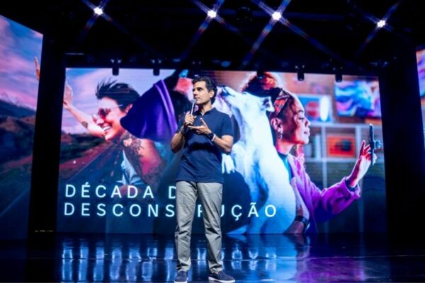 Um homem de meia-idade, vestindo uma camisa polo azul e calças claras, está em pé no palco com um microfone na mão, falando para a audiência. Atrás dele, um grande ecrã LED exibe imagens vibrantes de pessoas em movimento e a frase “DÉCADA DA DESCONSTRUÇÃO”.