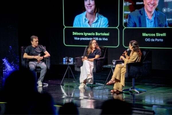 Três pessoas estão sentadas em cadeiras pretas num palco, participando de um painel de discussão. Há duas mulheres e um homem, todos com microfones. No ecrã atrás deles aparecem os nomes “Débora Ignacio Bortolasi” e “Marcos Sirelli”, com os respetivos cargos.