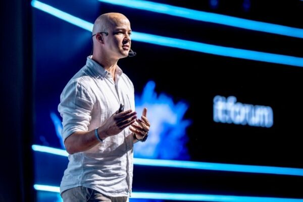 Um homem careca, com camisa branca de linho e microfone de cabeça, gesticula enquanto fala num palco iluminado por luzes azuis. Ao fundo, vê-se parcialmente a palavra "itforum" num ecrã.