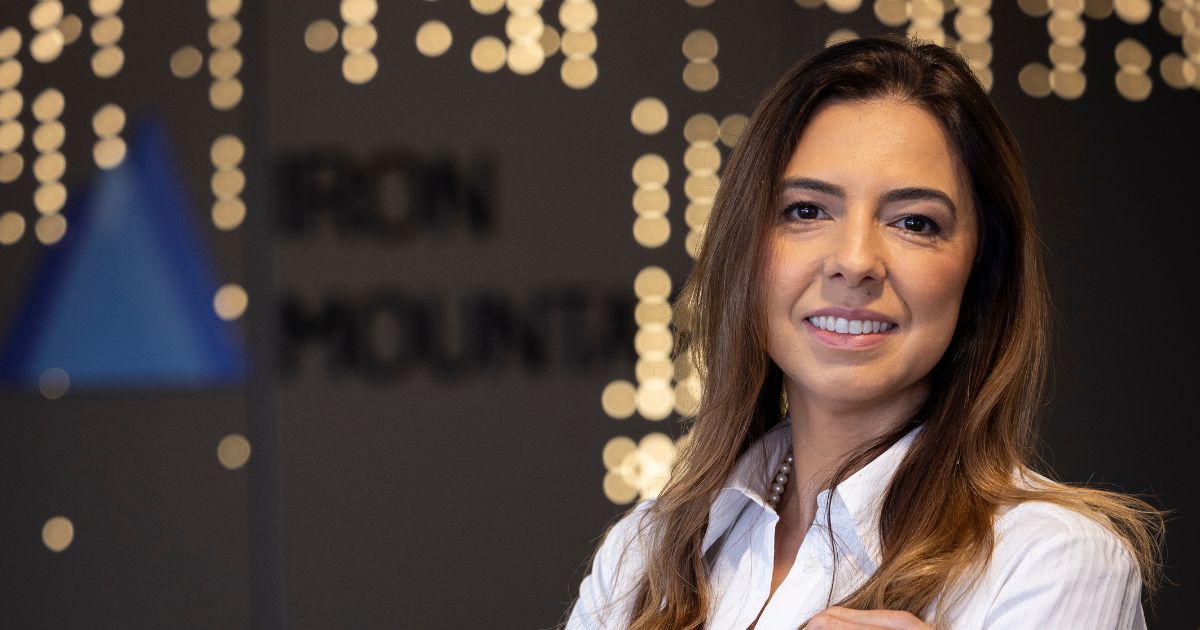 Ana Carla Martins Netto, diretora-geral comercial da Iron Mountain Brasil, sorri em frente ao logotipo da empresa. Ela veste camisa branca e colar de pérolas, com luzes desfocadas ao fundo criando um ambiente moderno e acolhedor.