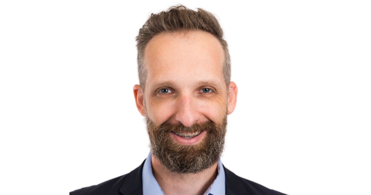 Homem branco de barba cheia e cabelos castanhos curtos, usando aparelho ortodôntico nos dentes, camisa azul clara e paletó escuro, posando contra fundo branco. Na imagem, está Ivo Bernard Schotten, novo CTO da SCforce.