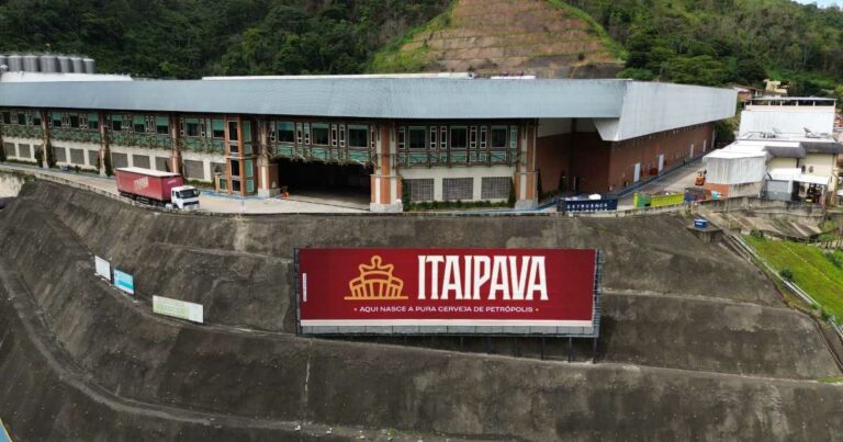 Imagem aérea da fábrica da cervejaria Itaipava, localizada em Petrópolis, Rio de Janeiro. Em destaque, há um grande letreiro vermelho com o logotipo da marca Itaipava e a frase "Aqui nasce a pura cerveja de Petrópolis". O edifício industrial apresenta arquitetura com estrutura metálica e paredes em alvenaria, rodeado por vegetação e áreas de encosta. Um caminhão com container também com a marca Itaipava está estacionado próximo à entrada da fábrica (alunos)