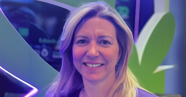 Helena Fragomeni, CEO da Hands-on, sorrindo em frente a um fundo colorido com elementos em verde, roxo e azul, possivelmente em um evento ou estande de tecnologia. A iluminação em tons roxos e azulados destaca seu rosto e cabelo loiro