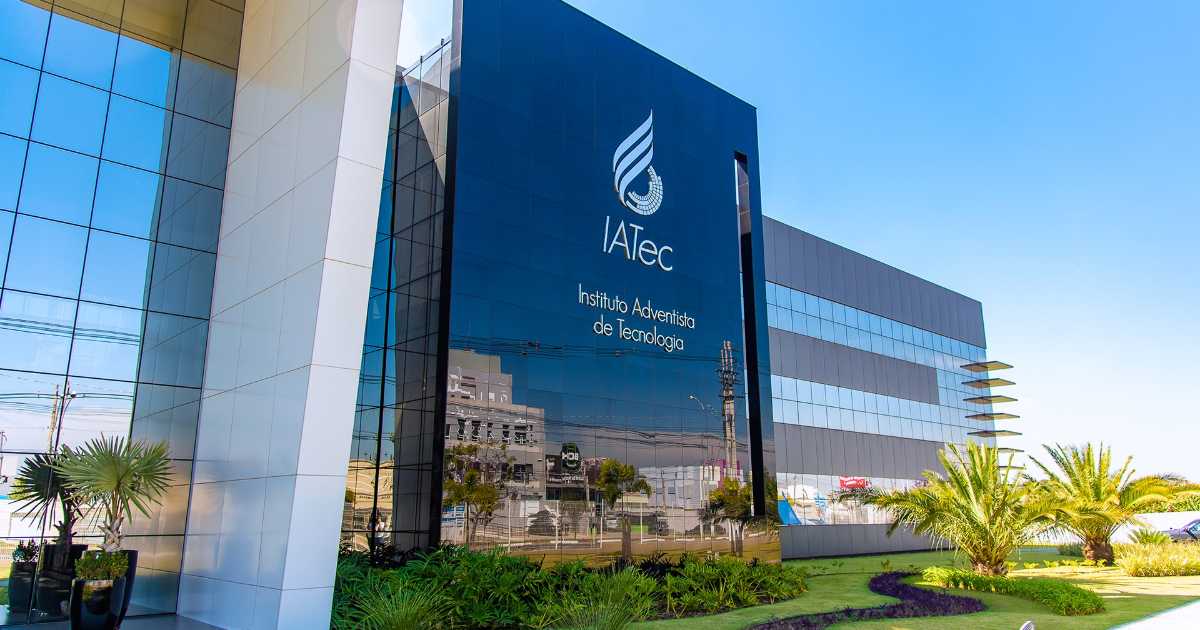 Prédio moderno com fachada espelhada e letreiro indicando "IATec – Instituto Adventista de Tecnologia", localizado em área urbana com paisagismo na frente, incluindo palmeiras, gramado e arbustos bem cuidados