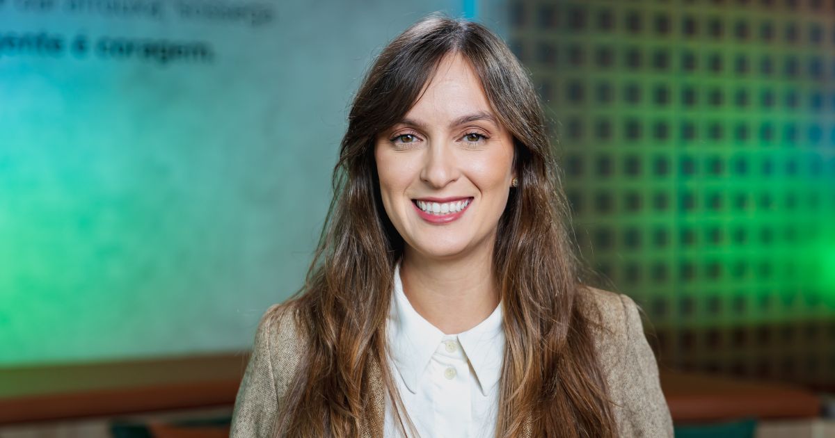 "Mulher jovem com cabelo castanho e longo, sorridente, vestida de forma profissional com camisa branca e blazer bege, posando num ambiente interior com fundo desfocado em tons verde-azulados." (JusBrasil)