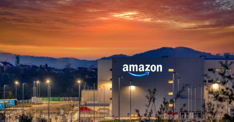 Centro logístico da Amazon ao entardecer, com o logótipo da empresa iluminado na fachada de um grande edifício industrial cinzento. O céu exibe tons vibrantes de laranja e vermelho ao pôr do sol, com colinas escuras e silhuetas de árvores ao fundo. À frente do armazém, há postes de iluminação acesos e contentores coloridos, sugerindo uma zona de carga e descarga. O ambiente mistura a grandiosidade do cenário natural com a modernidade das instalações da empresa.