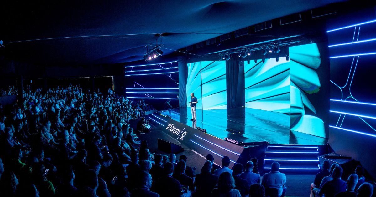 Auditório escuro e moderno, lotado de pessoas sentadas e atentas a uma apresentação num palco iluminado. Um orador está em pé no centro do palco, falando ao público. O cenário é envolvente, com luzes de LED azuis nas laterais e um grande ecrã ao fundo a exibir imagens abstratas em tons de azul e verde. As palavras "itforum" estão visíveis no palco, indicando que o evento faz parte do IT Forum.