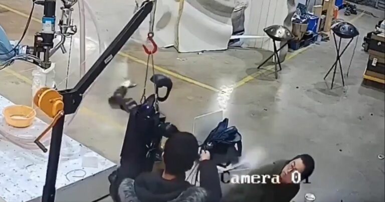 Fotograma de uma gravação de segurança (indicada pela legenda “Camera 01”) mostra uma mulher a ser atingida no rosto por um jato de líquido, aparentemente lançado acidentalmente por um braço robótico ou máquina industrial. Ao fundo vê-se um ambiente de oficina ou fábrica, com chão de cimento, ferramentas e equipamentos diversos.