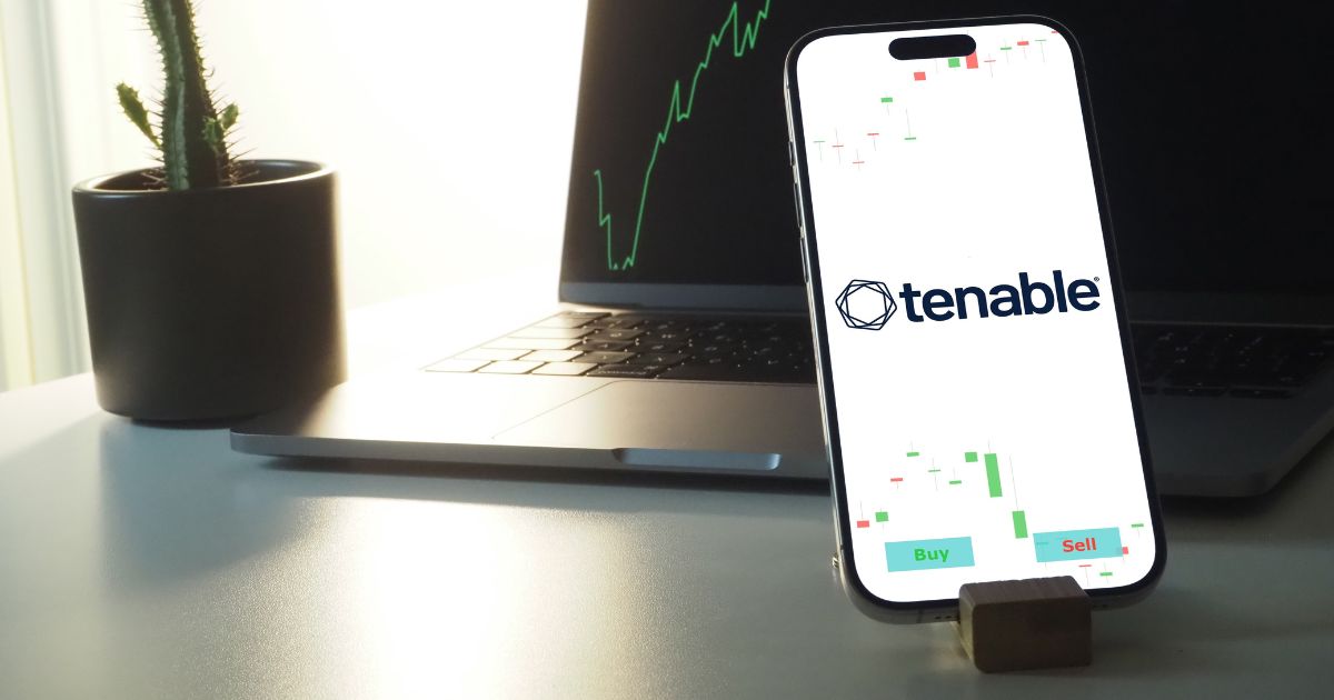 Um smartphone exibe o logotipo da Tenable e dados do mercado de ações, com botões "Buy" (comprar) e "Sell" (vender) na parte inferior da tela. O aparelho está apoiado em um suporte de madeira diante de um notebook aberto, que mostra um gráfico de linha verde. À esquerda do notebook, há um pequeno cacto decorativo.