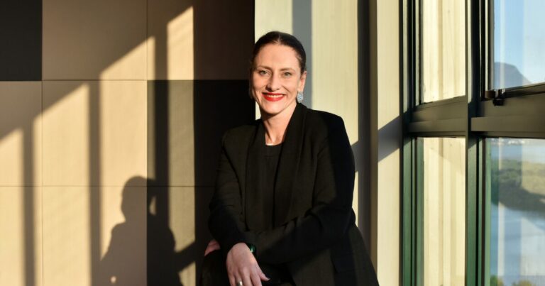 Auana Mattar, CIO da TIM Brasil, em um retrato profissional. Ela tem cabelos cacheados curtos e usa brincos pequenos. Está sorrindo levemente, com um fundo desfocado que mostra uma paisagem urbana e o céu azul