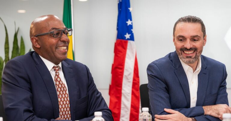 Dois homens bem vestidos, sentados lado a lado numa reunião, sorriem enquanto conversam. Ao fundo, vêem-se as bandeiras do Brasil e dos Estados Unidos, sugerindo um encontro diplomático ou empresarial entre os dois países.