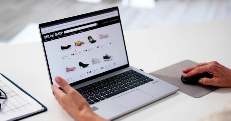Uma pessoa utiliza um computador portátil para navegar numa loja virtual de calçado. O ecrã mostra vários modelos de sapatos organizados em grelha, com preços e opções de cores. A mão direita está sobre um rato e a esquerda está próxima do teclado. Ao lado do portátil há uma folha com uma grelha e uns óculos (eset, mães)