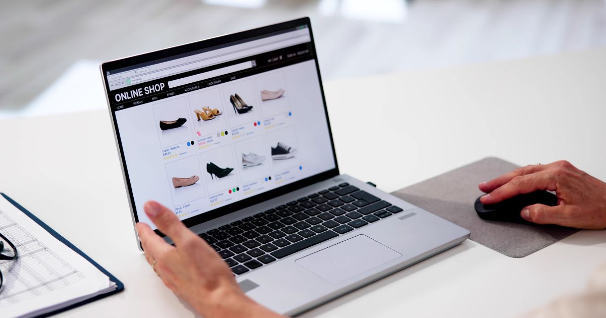 Uma pessoa utiliza um computador portátil para navegar numa loja virtual de calçado. O ecrã mostra vários modelos de sapatos organizados em grelha, com preços e opções de cores. A mão direita está sobre um rato e a esquerda está próxima do teclado. Ao lado do portátil há uma folha com uma grelha e uns óculos (eset, mães)
