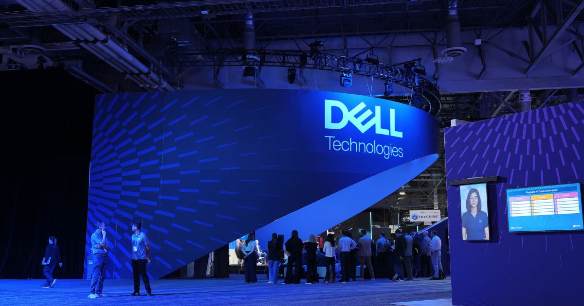 Grande stand da Dell Technologies numa feira tecnológica, iluminado em tons de azul. O logótipo da Dell Technologies está destacado numa parede curva e moderna, com várias pessoas a interagir e circular pelo espaço. À direita, vê-se um ecrã digital com informações e a imagem de uma colaboradora. (Nvidia)
