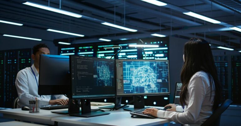Dois profissionais trabalham em um centro de dados moderno, sentados diante de computadores com múltiplos monitores. A mulher em primeiro plano observa telas que exibem representações gráficas de um cérebro digital e códigos de programação. O homem ao fundo também está concentrado em seu computador. O ambiente é escuro, iluminado pelas luzes dos equipamentos e servidores ao fundo, transmitindo uma atmosfera de alta tecnologia (Jitterbit )