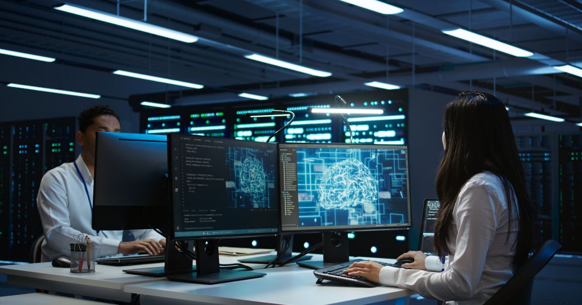 Dois profissionais trabalham em um centro de dados moderno, sentados diante de computadores com múltiplos monitores. A mulher em primeiro plano observa telas que exibem representações gráficas de um cérebro digital e códigos de programação. O homem ao fundo também está concentrado em seu computador. O ambiente é escuro, iluminado pelas luzes dos equipamentos e servidores ao fundo, transmitindo uma atmosfera de alta tecnologia (Jitterbit )