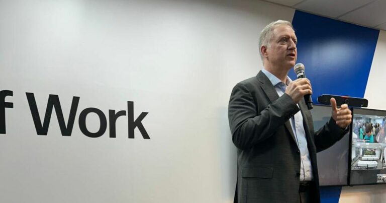 Um homem de fato escuro fala ao microfone em frente a uma parede branca com a inscrição parcial "of Work". Atrás dele, há um monitor a exibir imagens de câmaras de vigilância ou transmissão de uma reunião. O ambiente sugere uma apresentação ou conferência empresarial sobre o futuro do trabalho.