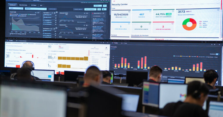 Imagem de um Centro de Operações de Segurança (SOC) da empresa Tivit, com vários profissionais concentrados em suas estações de trabalho, todos voltados para grandes painéis de monitoramento na parede. Os painéis exibem dashboards com gráficos, indicadores de segurança cibernética, tráfego de e-mails, vulnerabilidades detectadas e resultados de compliance. A imagem transmite um ambiente de vigilância tecnológica intensa, voltado à proteção de dados e gerenciamento de ameaças digitais. (Cibersegurança)
