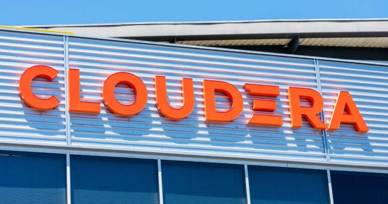 Fachada de prédio com o logotipo da empresa Cloudera em letras grandes e laranja, fixado em uma estrutura metálica prateada. A imagem foi tirada durante o dia, com céu azul ao fundo e janelas espelhadas na parte inferior da fachada