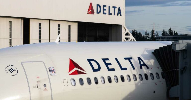 A imagem mostra um avião da companhia aérea Delta Air Lines estacionado em um portão de embarque, com parte do finger (ponte de embarque) acoplado à aeronave. Em destaque, vê-se o logotipo da Delta na lateral do avião e, ao fundo, um hangar branco com o mesmo logotipo e nome da empresa. À esquerda, aparece também o símbolo da aliança aérea SkyTeam, da qual a Delta faz parte. O cenário ao fundo inclui árvores e uma leve cobertura de nuvens no céu.