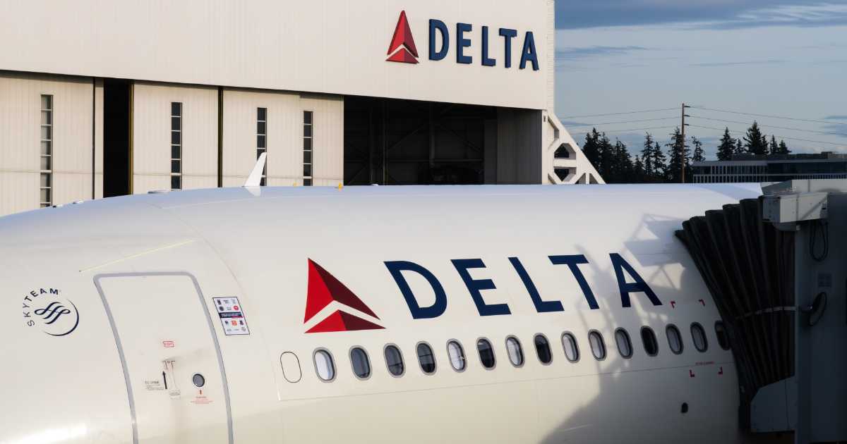 A imagem mostra um avião da companhia aérea Delta Air Lines estacionado em um portão de embarque, com parte do finger (ponte de embarque) acoplado à aeronave. Em destaque, vê-se o logotipo da Delta na lateral do avião e, ao fundo, um hangar branco com o mesmo logotipo e nome da empresa. À esquerda, aparece também o símbolo da aliança aérea SkyTeam, da qual a Delta faz parte. O cenário ao fundo inclui árvores e uma leve cobertura de nuvens no céu.