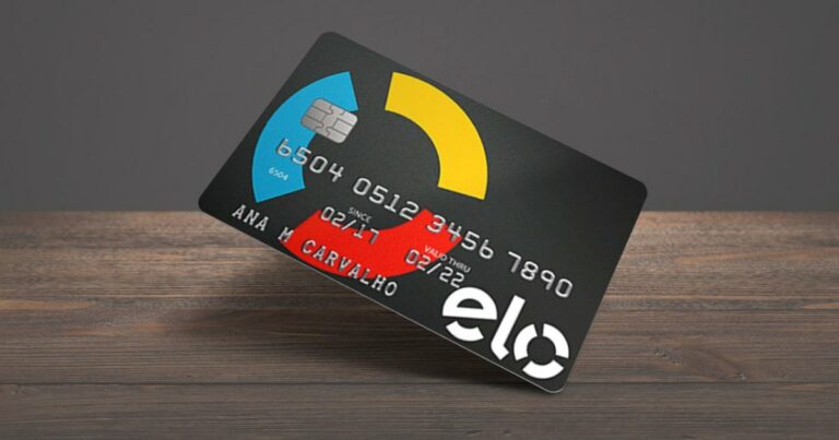 Cartão de crédito da bandeira Elo com fundo preto e elementos gráficos nas cores azul, amarelo e vermelho. O cartão está inclinado sobre uma superfície de madeira. Ele exibe um chip, o número do cartão, datas de validade (02/17 a 02/22) e o nome da titular, Ana M. Carvalho. O logotipo da Elo aparece em branco no canto inferior direito (solução)