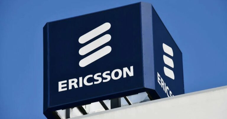 Placa com o logotipo da empresa Ericsson em destaque. O letreiro é azul-escuro com o nome "ERICSSON" em letras brancas, acompanhado do símbolo icônico da marca — três linhas brancas inclinadas paralelas. A placa está posicionada no topo de um edifício, com céu azul ao fundo (ações)