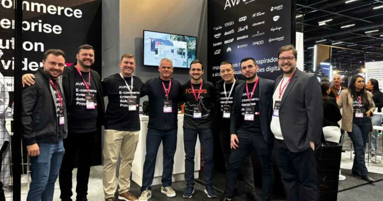 Imagem mostra os times da Avanti e FCamara reunidos em um evento de tecnologia, provavelmente voltado para e-commerce. O grupo é composto por nove homens, todos sorrindo e usando crachás de identificação. Muitos estão vestindo camisetas com as marcas "Avanti" ou "FCamara". Ao fundo, vê-se um estande com telas, banners promocionais e logotipos de diversas marcas, indicando um ambiente de feira ou congresso. O local é bem iluminado e moderno, com outros participantes visíveis ao fundo