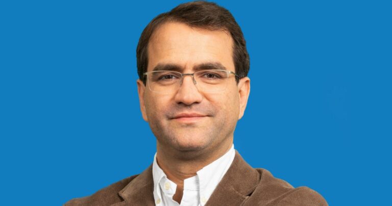 Bruno Domingues, head global de serviços financeiros da Intel, veste paletó marrom e camisa branca, posando com expressão serena diante de fundo azul sólido (IA generativa)