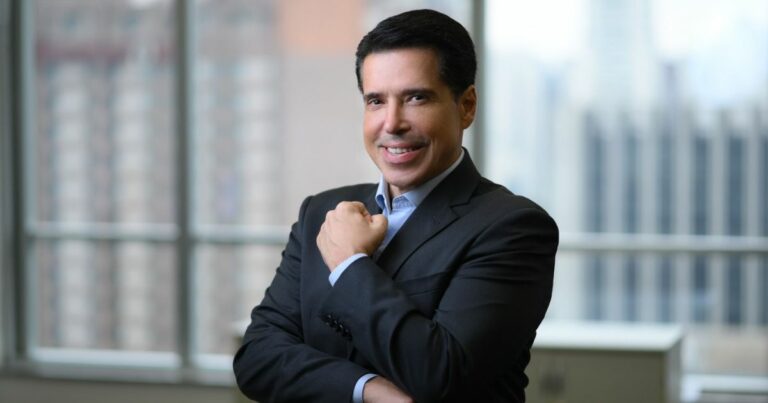 Carlos Perobelli, diretor-executivo da Nexmuv e idealizador da The Garage, posando sorridente, de terno escuro, em um ambiente corporativo com janelas amplas e vista desfocada de prédios ao fundo.