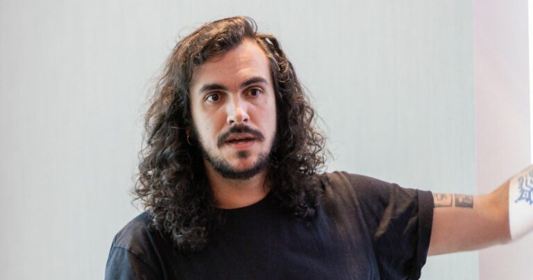 Lucas Bacic, CEO da Loja Integrada, em apresentação. Ele está com expressão atenta, usando camiseta preta e apontando para o lado, com tatuagens visíveis no braço e cabelos longos cacheados.