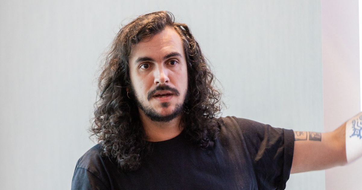 Lucas Bacic, CEO da Loja Integrada, em apresentação. Ele está com expressão atenta, usando camiseta preta e apontando para o lado, com tatuagens visíveis no braço e cabelos longos cacheados.