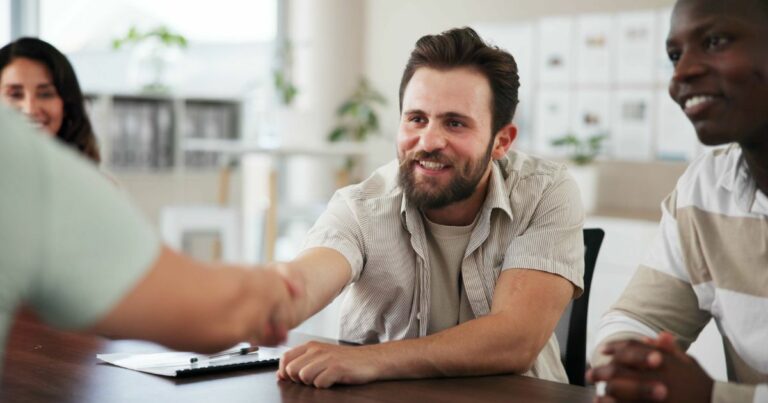 Um grupo de pessoas está sentado ao redor de uma mesa, em um ambiente que parece ser um escritório ou sala de reuniões. Uma pessoa estende a mão para cumprimentar outra, cuja mão aparece em primeiro plano. Ao fundo, há prateleiras e algumas plantas, compondo um cenário corporativo ou profissional. (Simetrik)