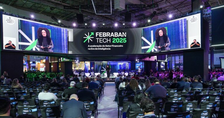 Auditório principal da Febraban Tech 2025, com público sentado assistindo a uma apresentação no palco. No centro do palco, uma mulher de cabelos cacheados e volumosos fala ao microfone, enquanto sua imagem é projetada em um grande telão curvo acima dela. O telão exibe também o logotipo do evento e o tema: "Febraban Tech 2025 – A aceleração do Setor Financeiro na Era da Inteligência". Nas laterais do telão, há intérpretes de Libras. O ambiente é moderno e tecnológico, com iluminação roxa e verde e estandes de empresas visíveis ao fundo. (5G, 6g)