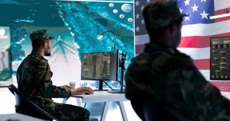 Dois militares em trajes de camuflagem estão sentados em frente a estações de trabalho com computadores, operando sistemas de monitoramento ou vigilância. Ao fundo, uma grande tela exibe uma imagem aérea em tons de azul, possivelmente de um drone ou satélite, e parte da bandeira dos Estados Unidos é visível no canto direito. A cena sugere um ambiente de operações militares de alta tecnologia, com ênfase em vigilância ou controle remoto. (Meta) (OpenAI)